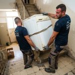 שני מובילים מקצועיים סוחבים בזהירות מכונת כביסה כבדה במורד חדר מדרגות צר בבניין מגורים ישן ללא מעלית.
