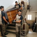 צוות מובילים מקצועי של חברת פינוי דירות מוריד בזהירות שידת עץ כבדה בגרם מדרגות של בניין מגורים ללא מעלית.